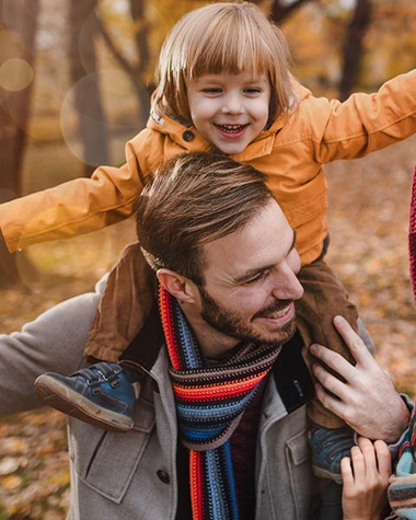 Das Bild zeigt eine Frau und einen Mann, die einen Jungen auf ihren Schultern tragen. Alle drei lächeln. Sie befinden sich in einem Wald mit herbstlicher Atmosphäre, auf dessen Boden viele Blätter liegen.