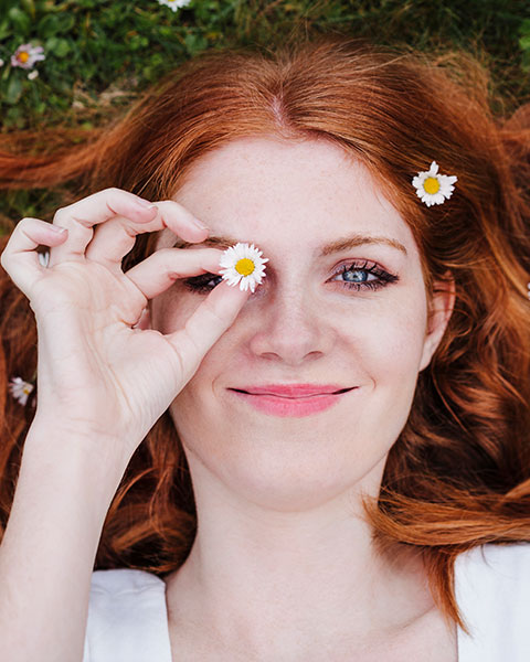 Eine Frau mit roten Haaren lächelt und hält eine Blume vor ihr Auge, während sie im Gras liegt.