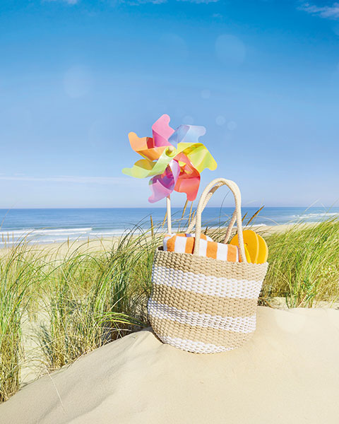  Eine Strandtasche mit einem bunten Windfänger steht auf einem Sandhaufen, im Hintergrund Strand, Pflanzen und das blaue Meer.
