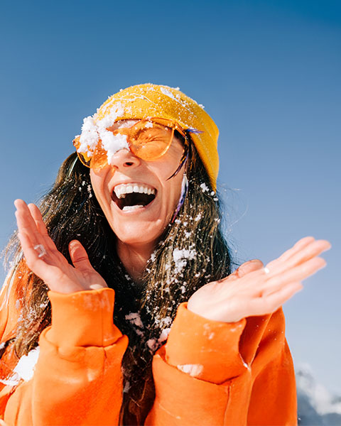 Eine Person lacht im Schnee und trägt eine orange Jacke sowie eine gelbe Mütze.