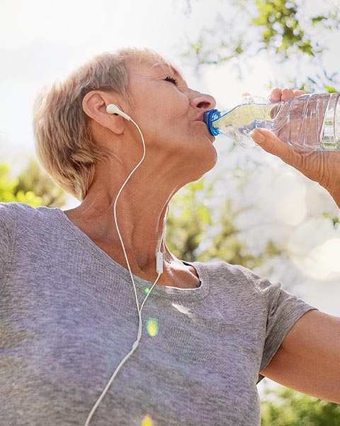 Eine joggende Frau trinkt Wasser aus einer Flasche in einer grünen, sonnigen Umgebung.